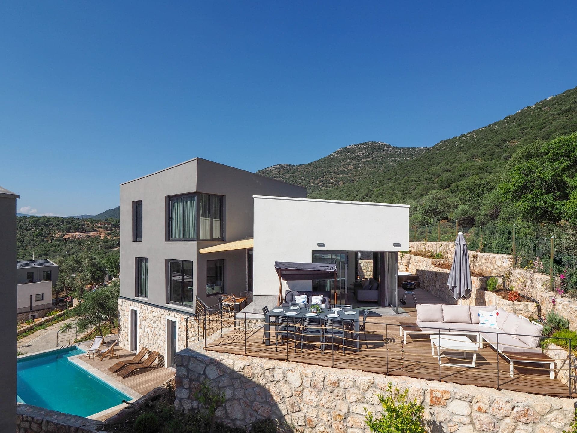 Ahşap teraslı yemek alanı ve oturma grubu olan havuzlu villa dış cephesi.