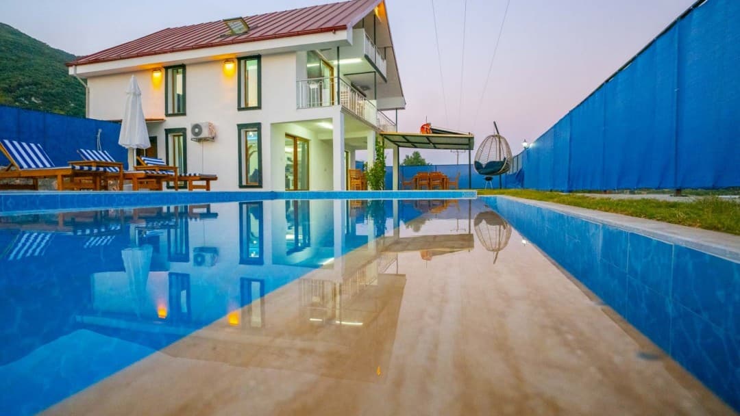 Havuz kenarında iki katlı beyaz villa, şezlonglar ve salıncak bulunan bahçe.