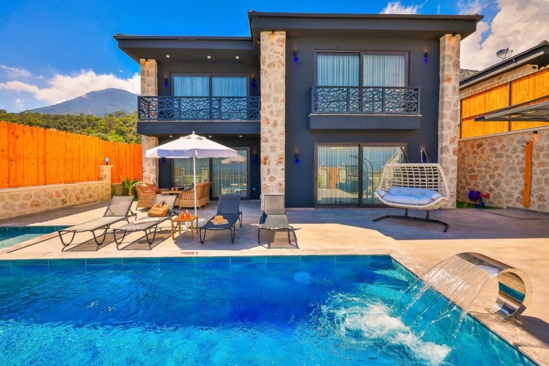 Şelale detaylı mavi havuz ve arkasında modern iki katlı koyu renk villa.