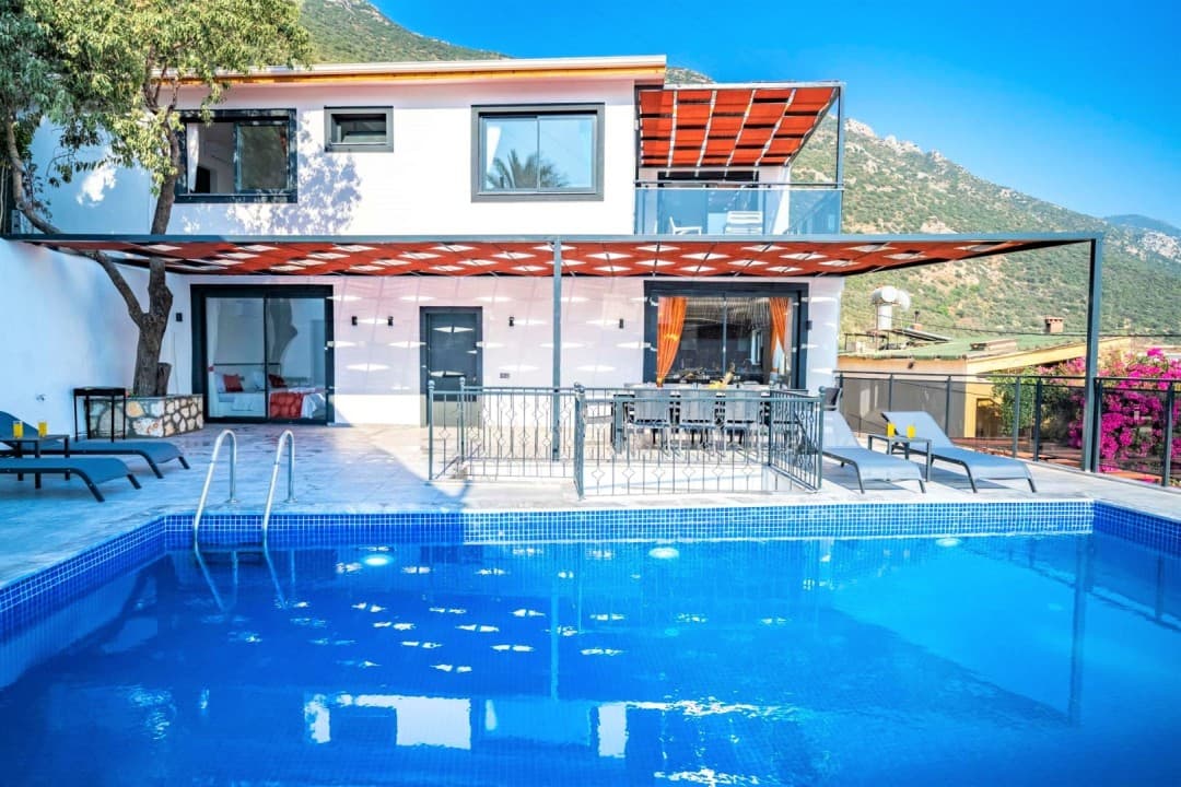 Dağ manzaralı, önünde mavi yüzme havuzu, şezlonglar ve gölgelikli oturma alanı bulunan beyaz iki katlı villa.