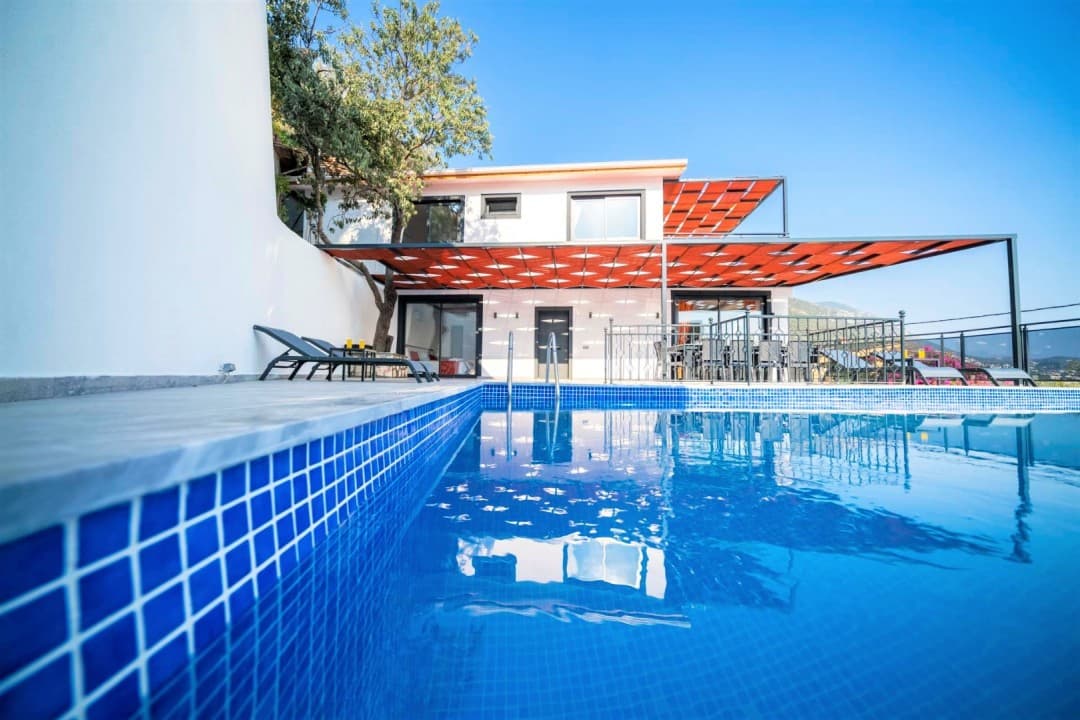 Açık mavi yüzme havuzunun arkasında turuncu gölgelikli beyaz iki katlı bir villa yer alıyor.