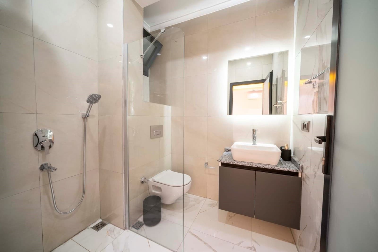 Bej seramik kaplı banyoda duş kabini, asma klozet ve ışıklı ayna bulunan lavabo yer alıyor.