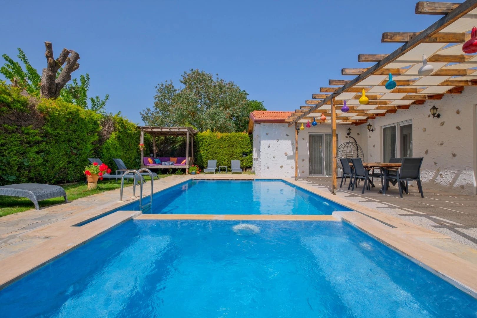 Mavi yüzme havuzu ve ahşap pergola altında yemek masası bulunan güneşli bahçe.