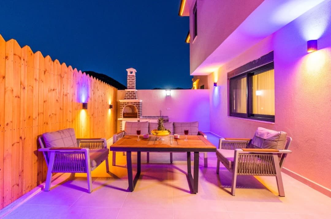 Gece aydınlatmalı, ahşap çitli terasta ahşap masa, gri koltuklar ve tuğla barbekü bulunan villa bahçesi.