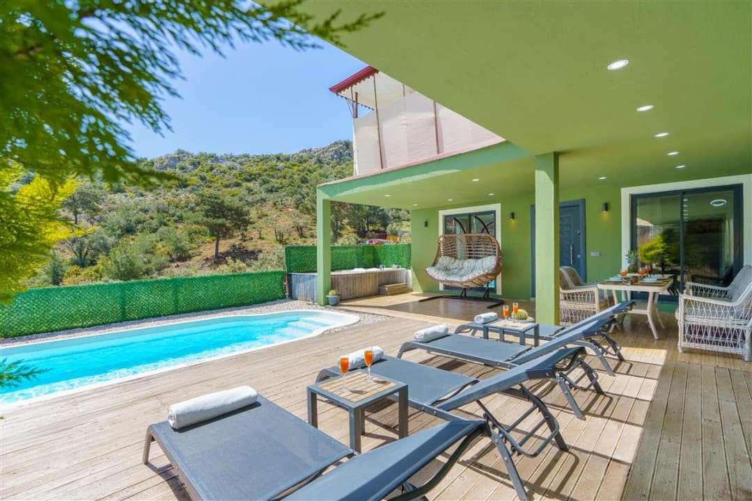 Ahşap zeminli terasta şezlonglar, havuz, salıncak ve dağ manzaralı yeşil cepheli bir villa.