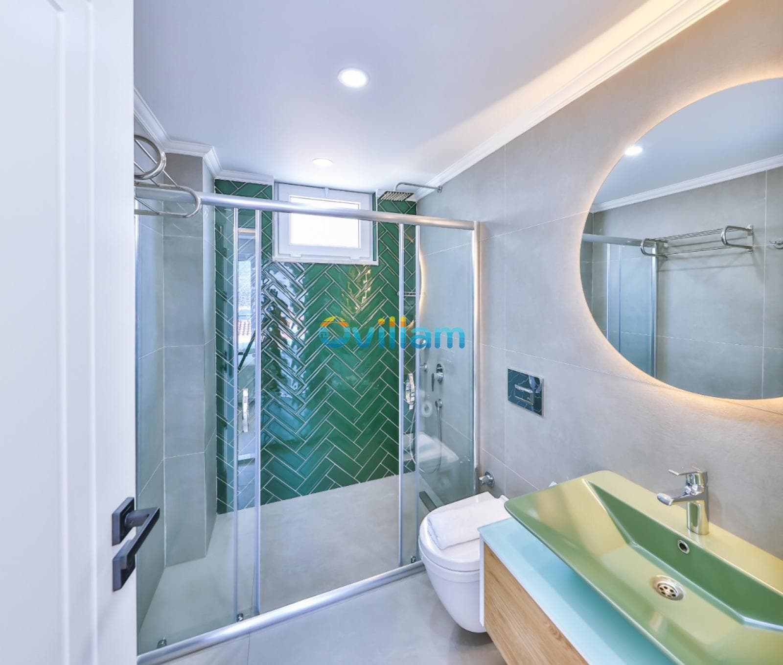 Yeşil seramik duvarlı ve duşakabinli modern banyoda yuvarlak ayna ve lavabo yer alıyor.
