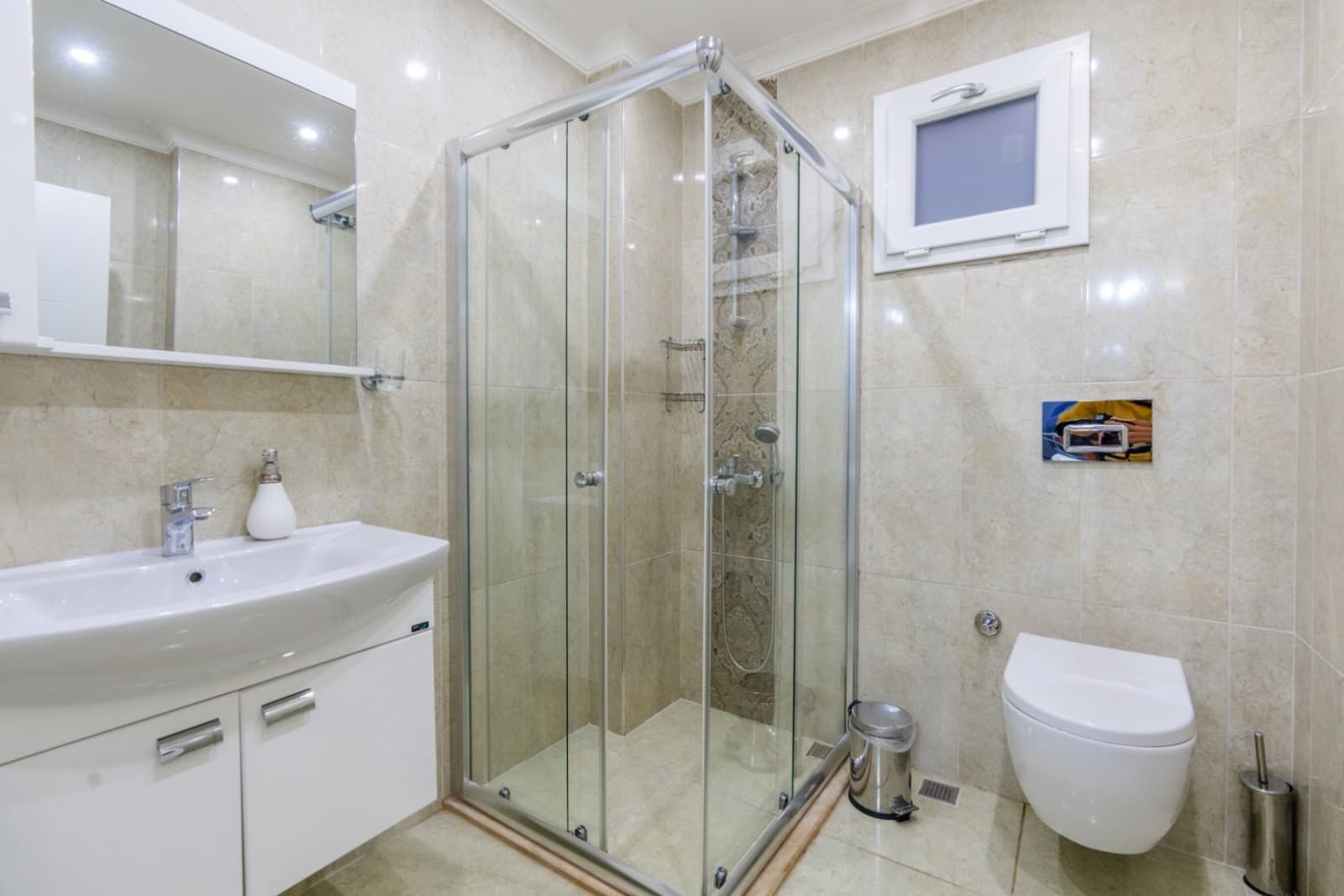 Bej seramik kaplı, cam duşakabinli ve beyaz lavabolu banyo.