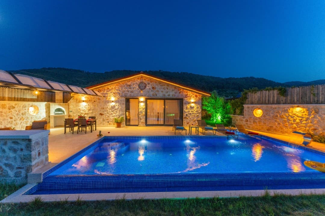 Gece aydınlatmalı taş duvarlı villa, önünde özel havuz, şezlonglar ve yemek masası bulunan geniş bir teras.