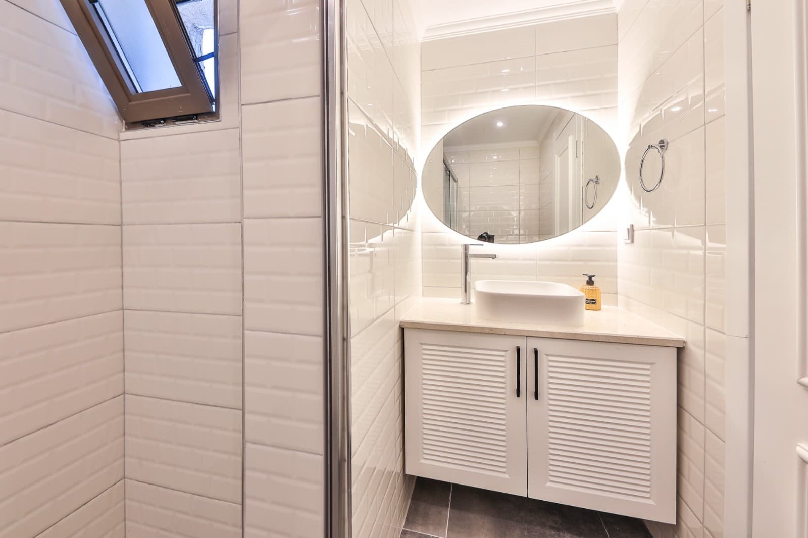 Beyaz seramikli banyoda oval aydınlatmalı ayna ve lavabo yer alıyor.