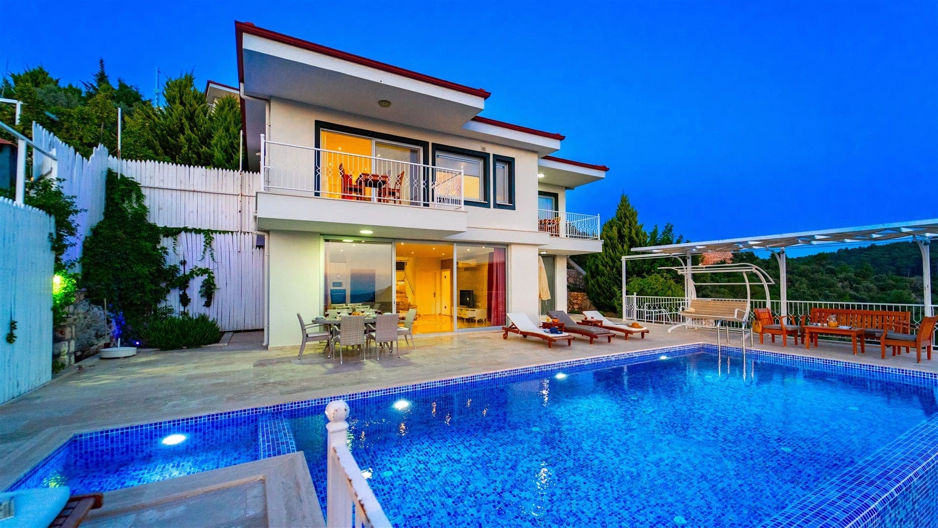 Akşam vakti aydınlatmalı havuz kenarında şezlonglar ve iki katlı villa yer alıyor.