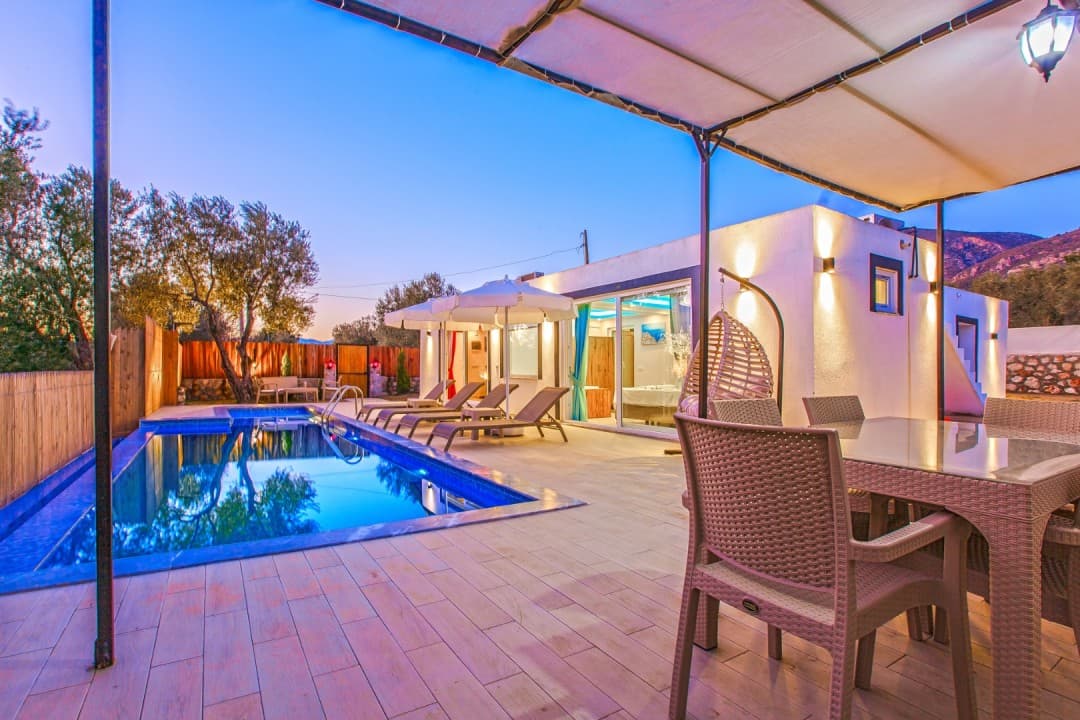 Akşam vaktinde havuz kenarında şezlonglar, yemek masası ve asılı salıncak bulunan modern villa bahçesi.
