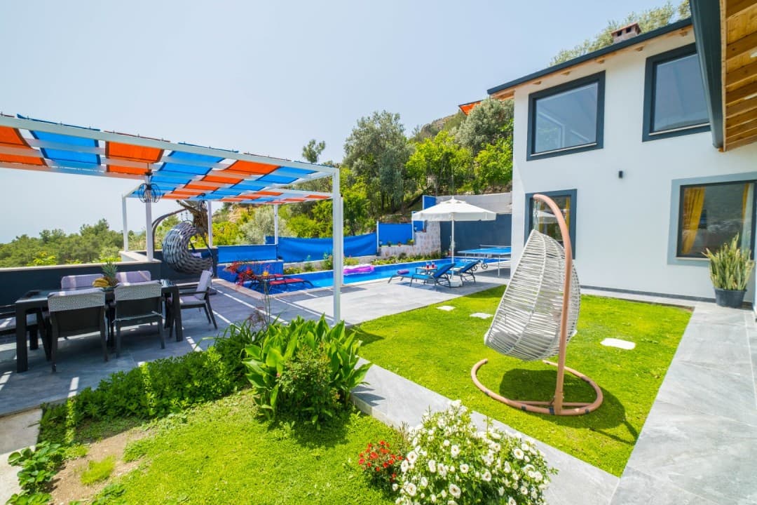 Bahçesinde havuz, şezlonglar, salıncak ve yemek masası olan beyaz dış cepheli villa.