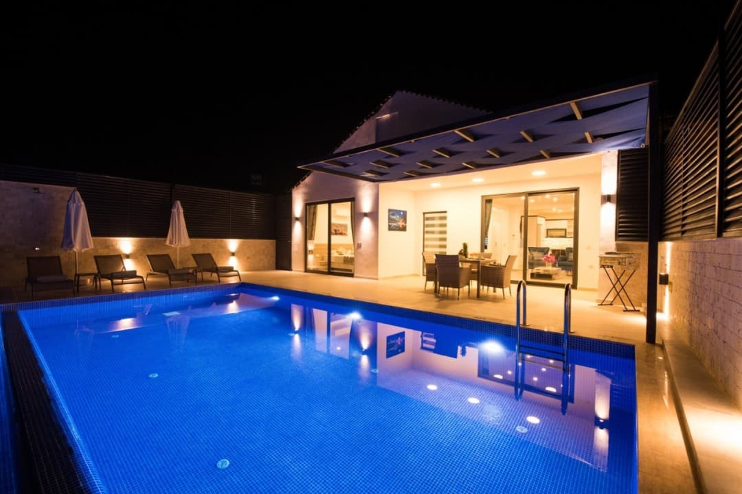 Gece aydınlatmalı havuzu, şezlongları ve veranda masası bulunan modern villanın dış görünümü.