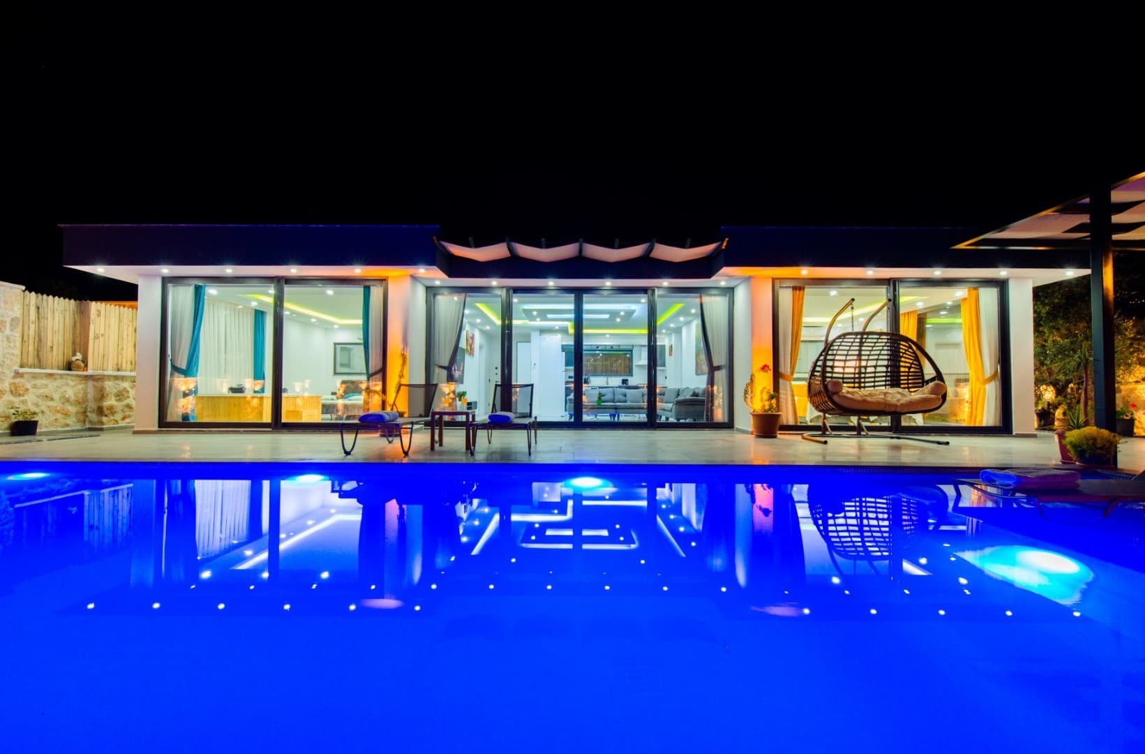 Gecenin karanlığında havuz aydınlatmalı modern villanın dış cephesi ve havuz kenarındaki oturma grubu.