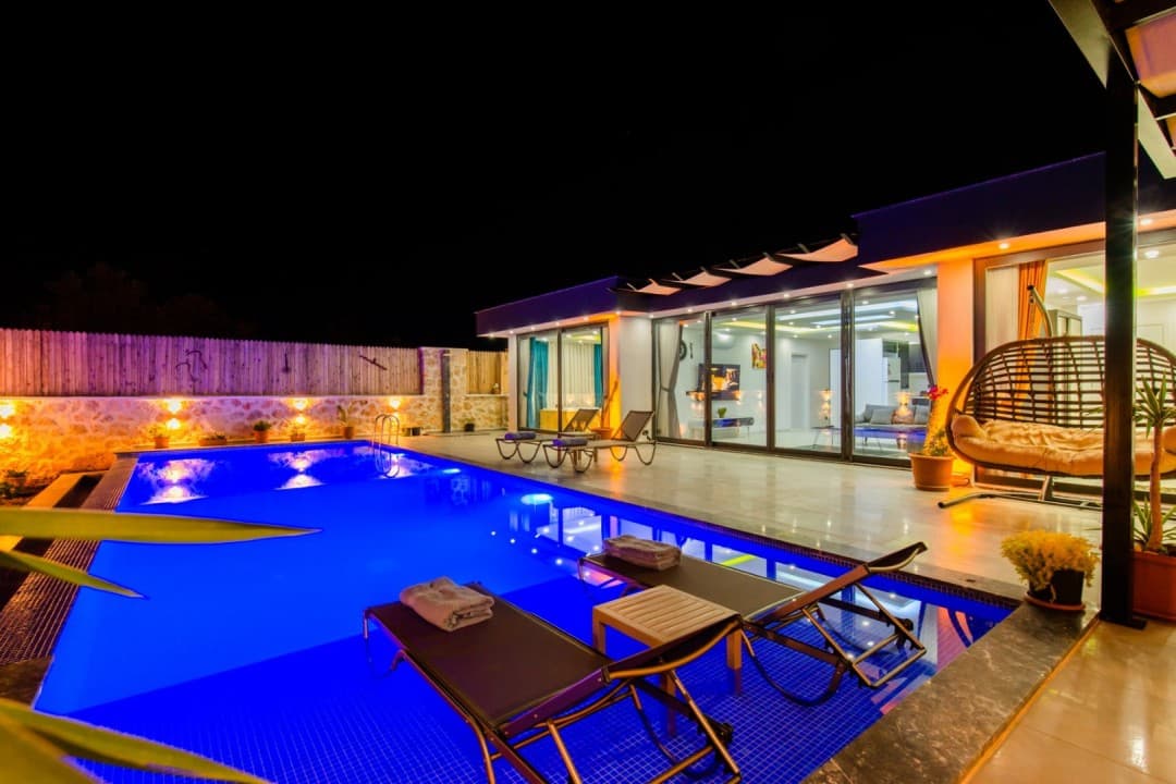 Gece aydınlatmalı havuz kenarında şezlonglar ve arka planda villa dış cephesi.