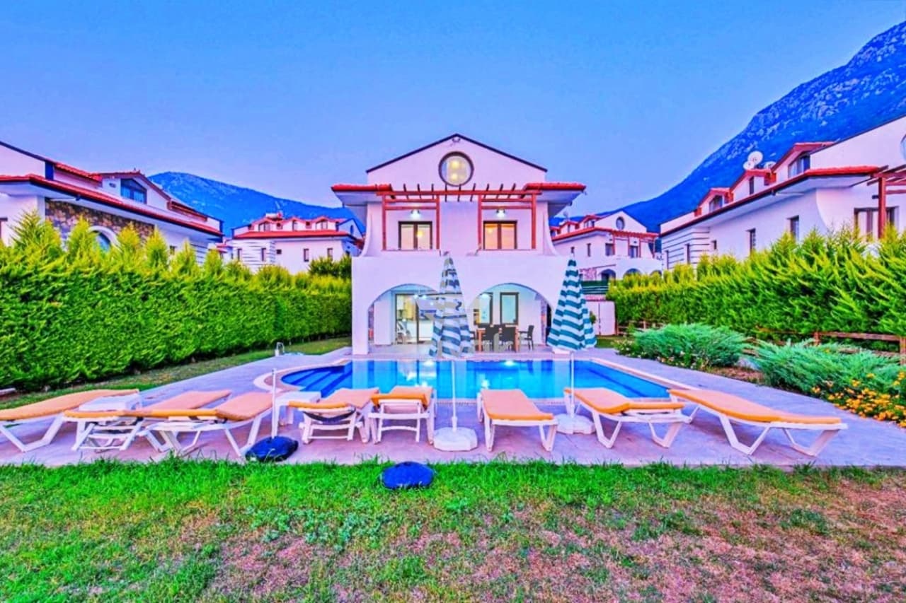 Önünde havuz ve şezlonglar bulunan beyaz iki katlı villa, arkasında yeşil çitler ve dağlar var.