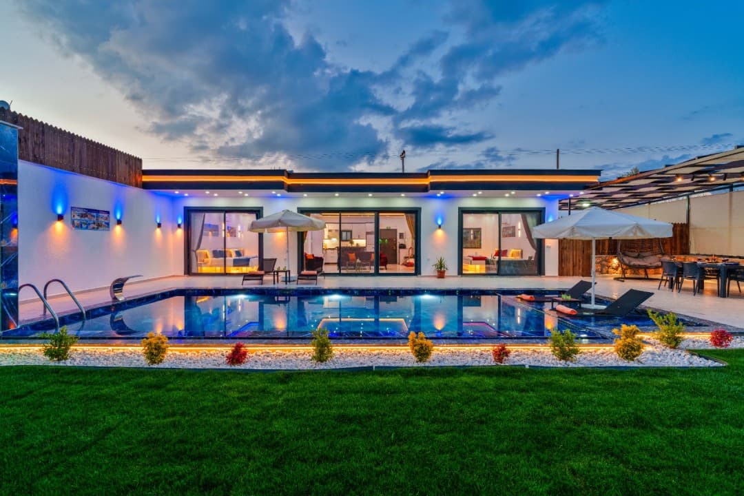 Akşam vakti aydınlatılmış havuzu, şezlongları ve çim bahçesi bulunan modern tek katlı villa.