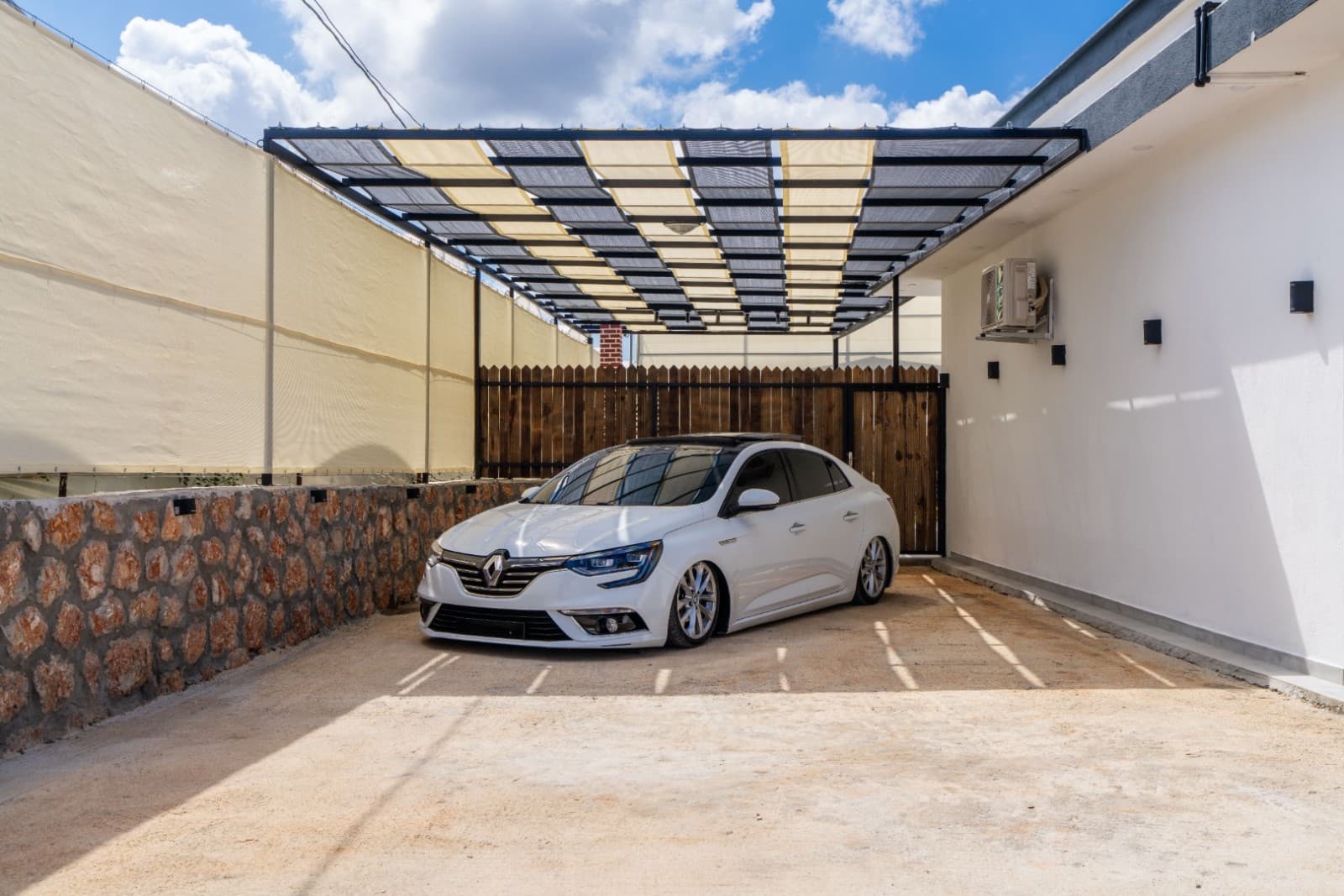 Ahşap ve taş duvarlı otopark alanında gölgelik altında park edilmiş beyaz otomobil.