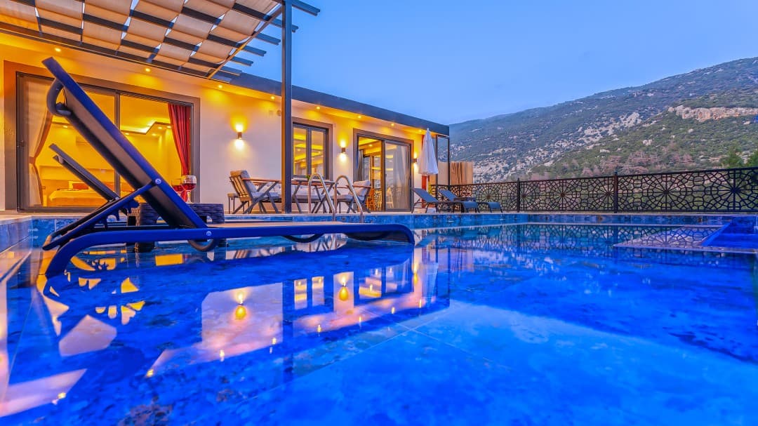 Akşam vakti aydınlatmalı havuz ve şezlonglar önünde dağ manzaralı tek katlı villa.