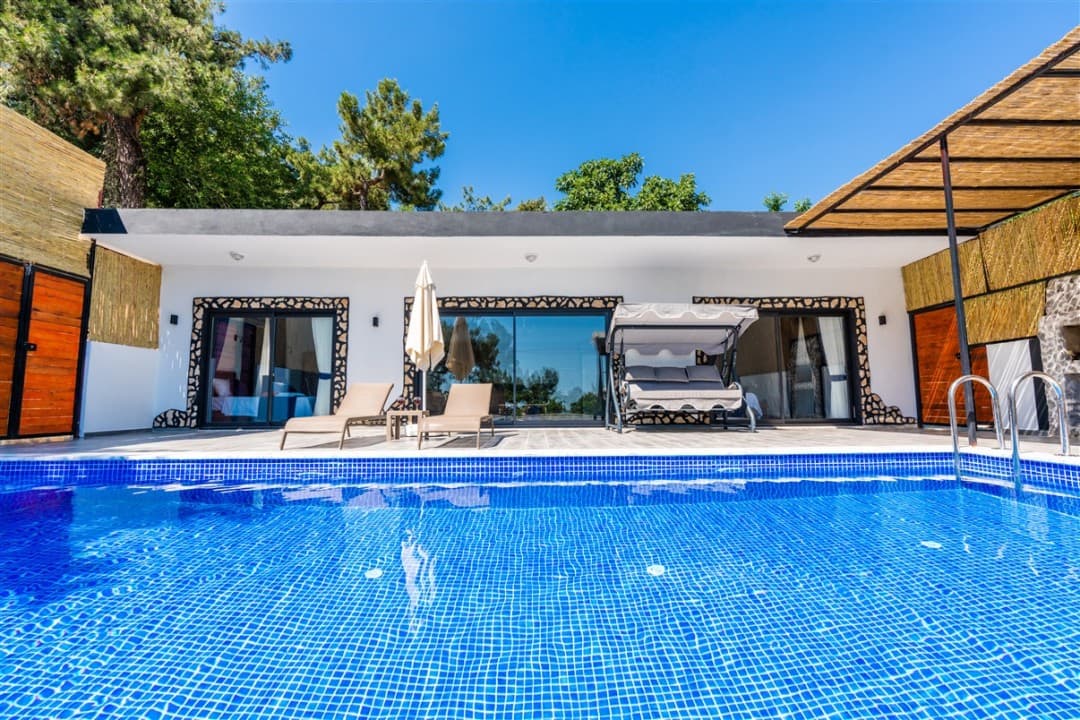 Gündüz mavisi havuz kenarında şezlonglar ve salıncak bulunan villa cephesi.