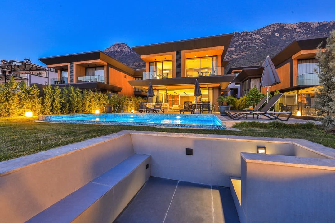 Işıklandırılmış havuzun arkasında modern çift katlı villa ve dağ silüeti.