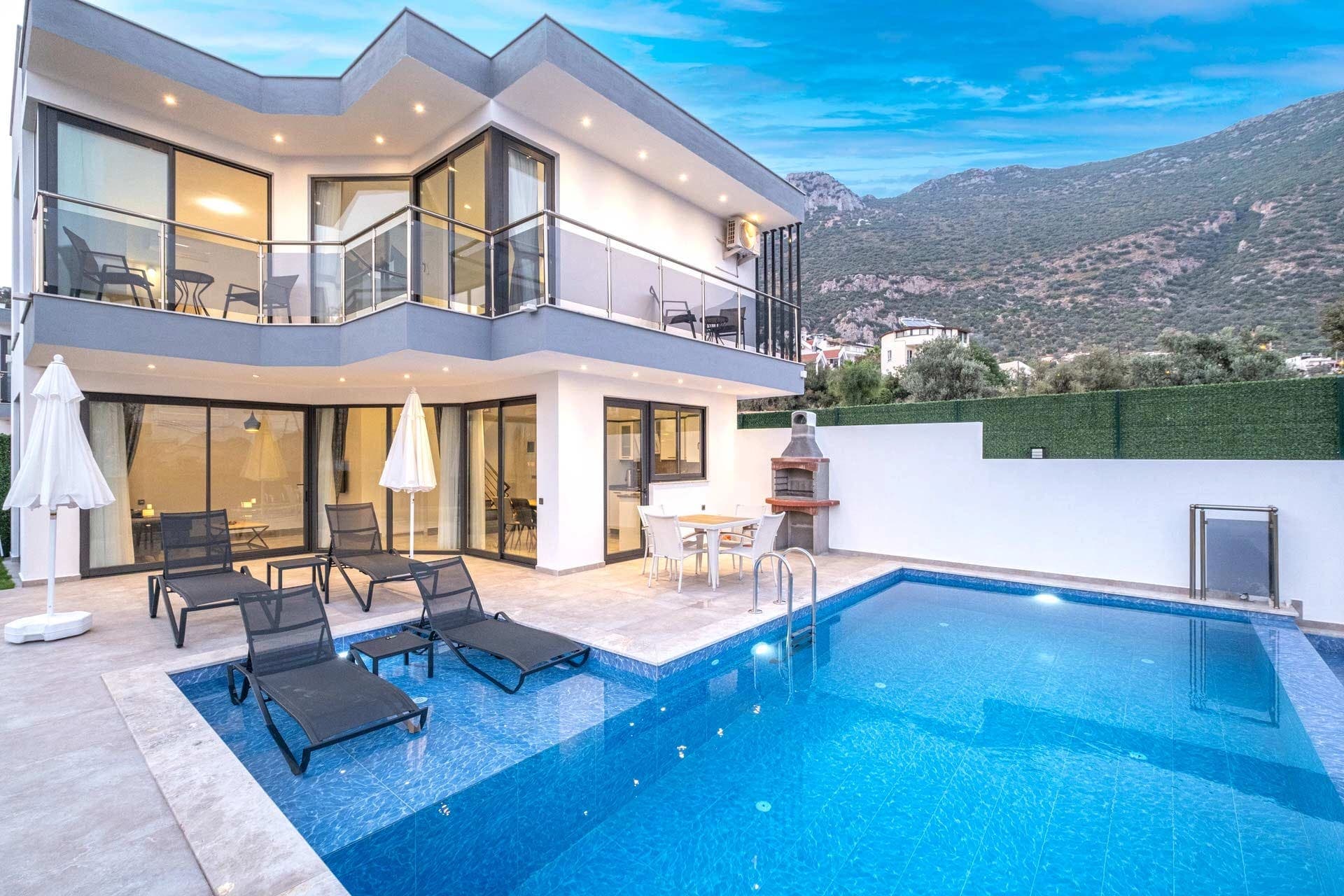 Dağ manzaralı havuz başında şezlonglar ve şemsiyeler bulunan villa dış cephesi.
