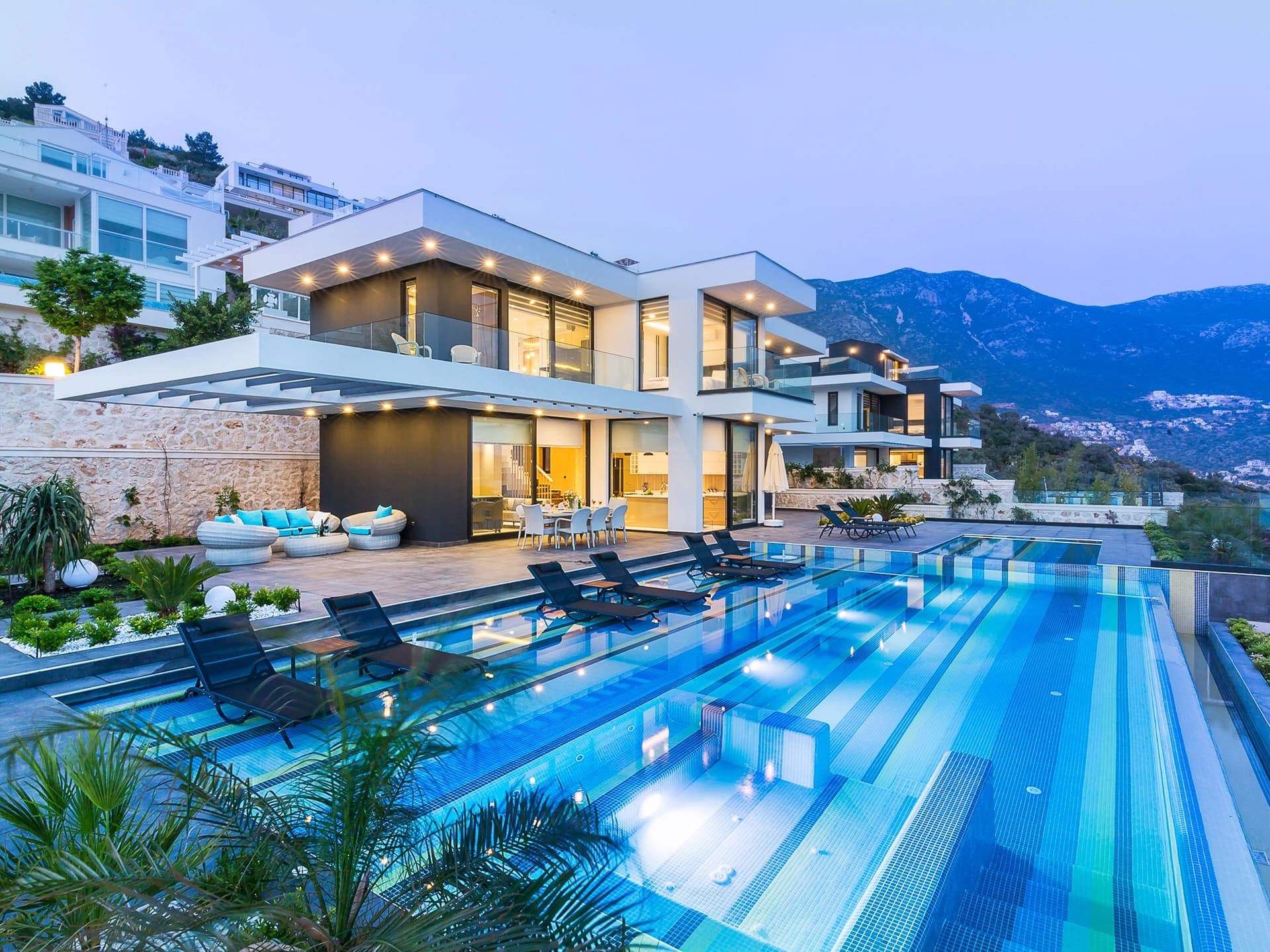 Akşam vakti aydınlatmalı havuzu ve dağ manzarası olan modern lüks villa dış cephesi.