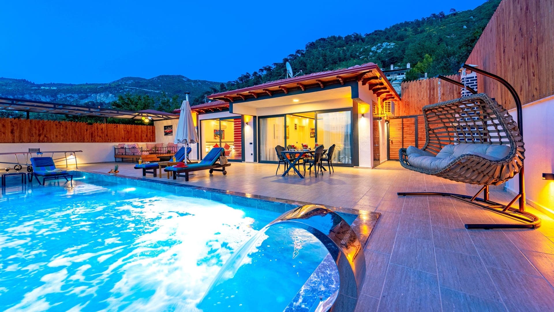 Gece aydınlatmalı havuz başı, salıncak koltuğu ve dağ manzaralı villa dış mekanı.