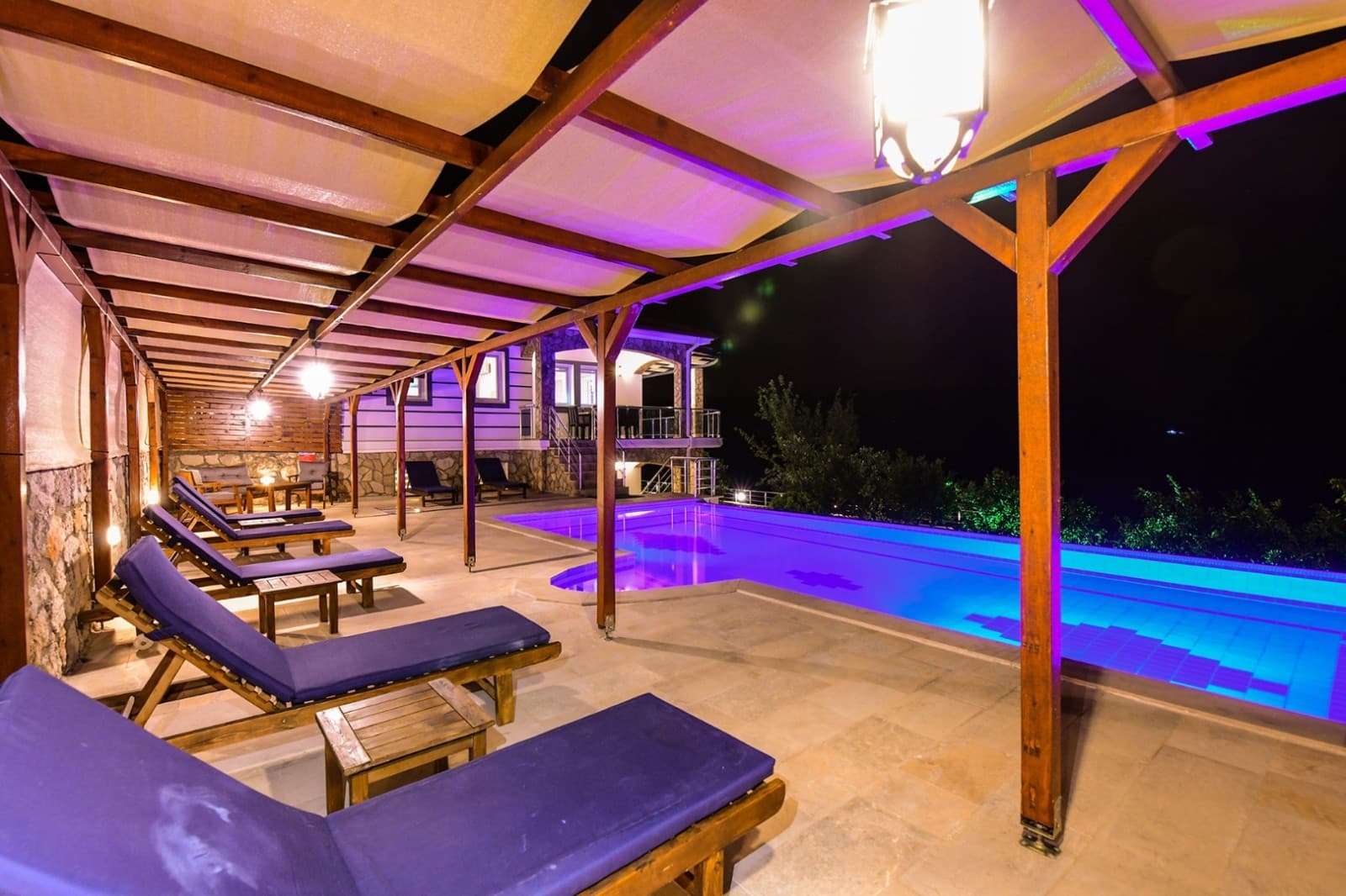 Ahşap pergola altında mor minderli şezlonglar ve aydınlatmalı havuzun gece görünümü.
