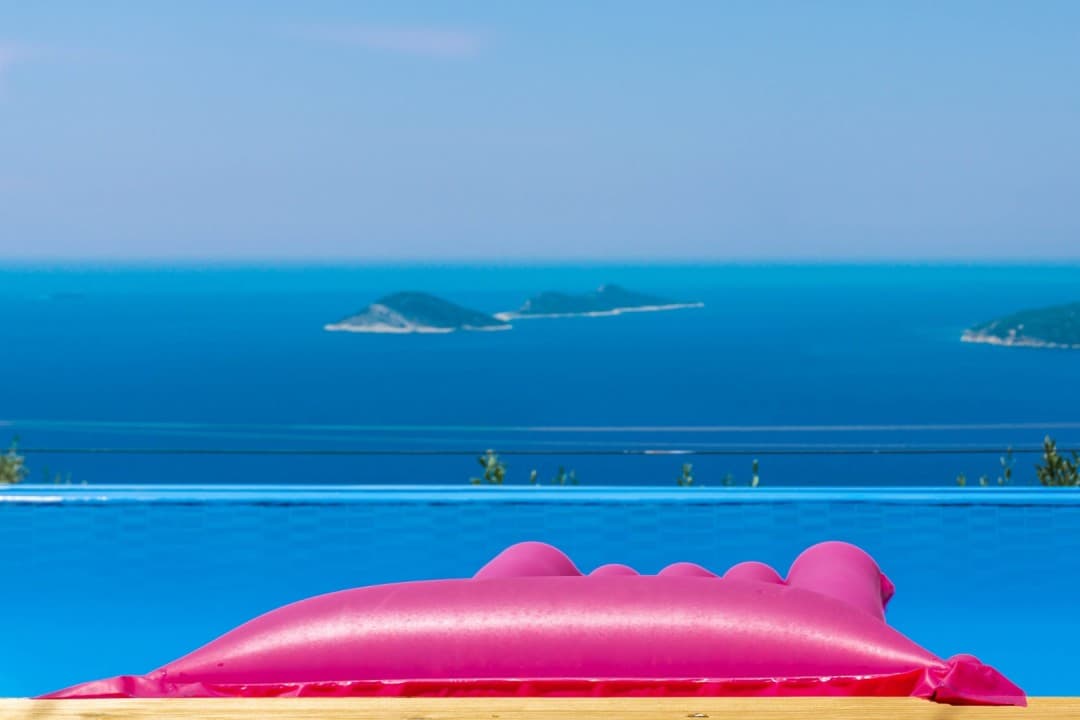 Havuz kenarında pembe şişme yatak ve arka planda deniz manzarası.