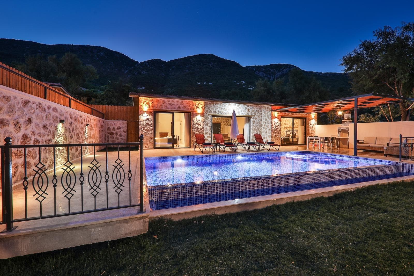 Gece aydınlatmalı taş duvarlı villa, önünde yüzme havuzu, şezlonglar ve dağ manzarası.