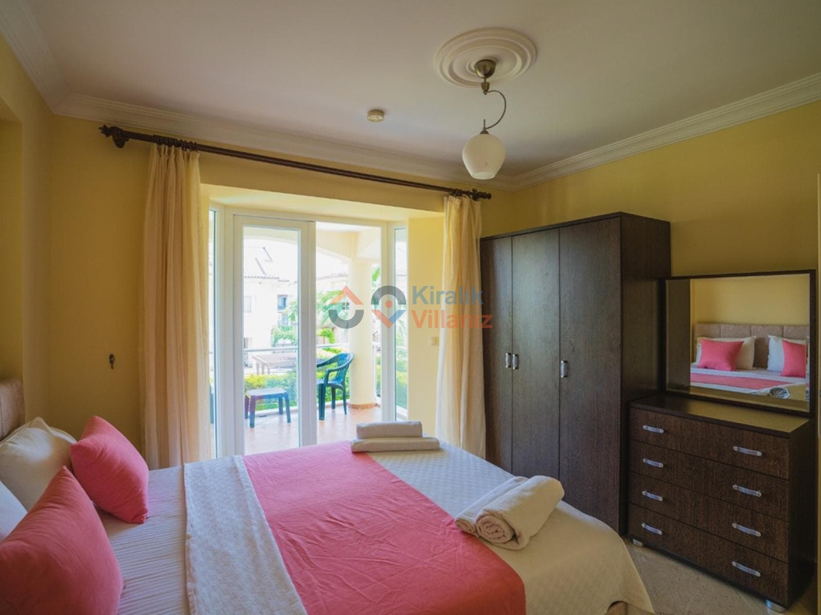 Pembe yatak örtülü çift kişilik yatak, ahşap gardırop ve balkon kapısı.