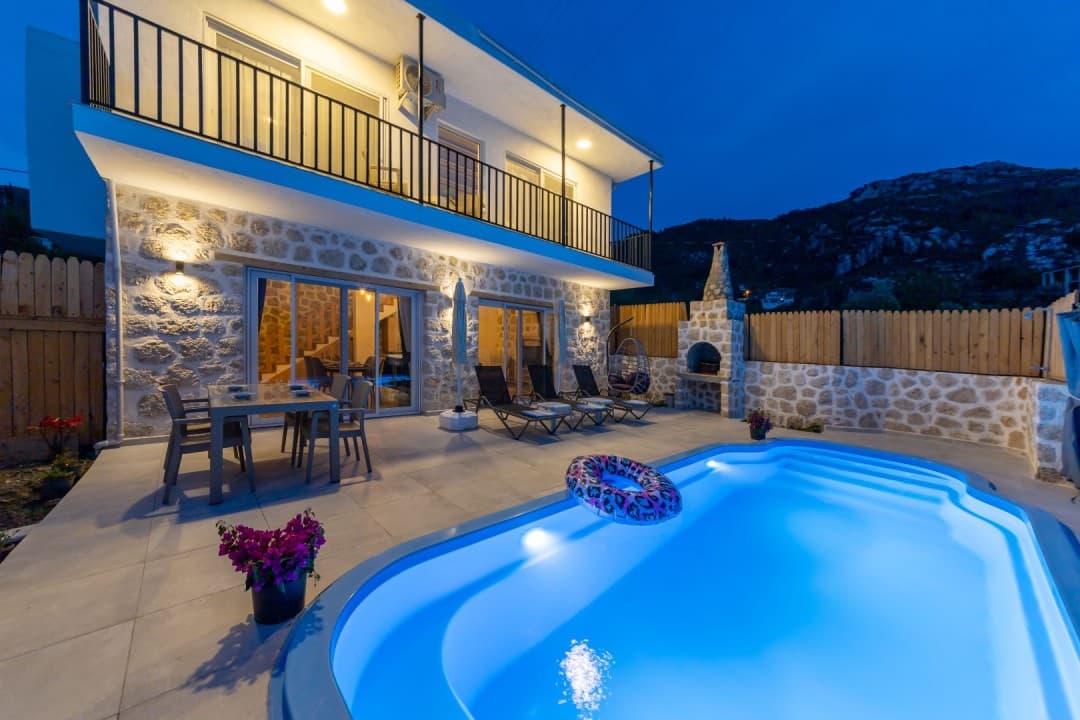 Akşam vakti aydınlatmalı havuzu, şezlongları ve yemek masası olan iki katlı taş villa.