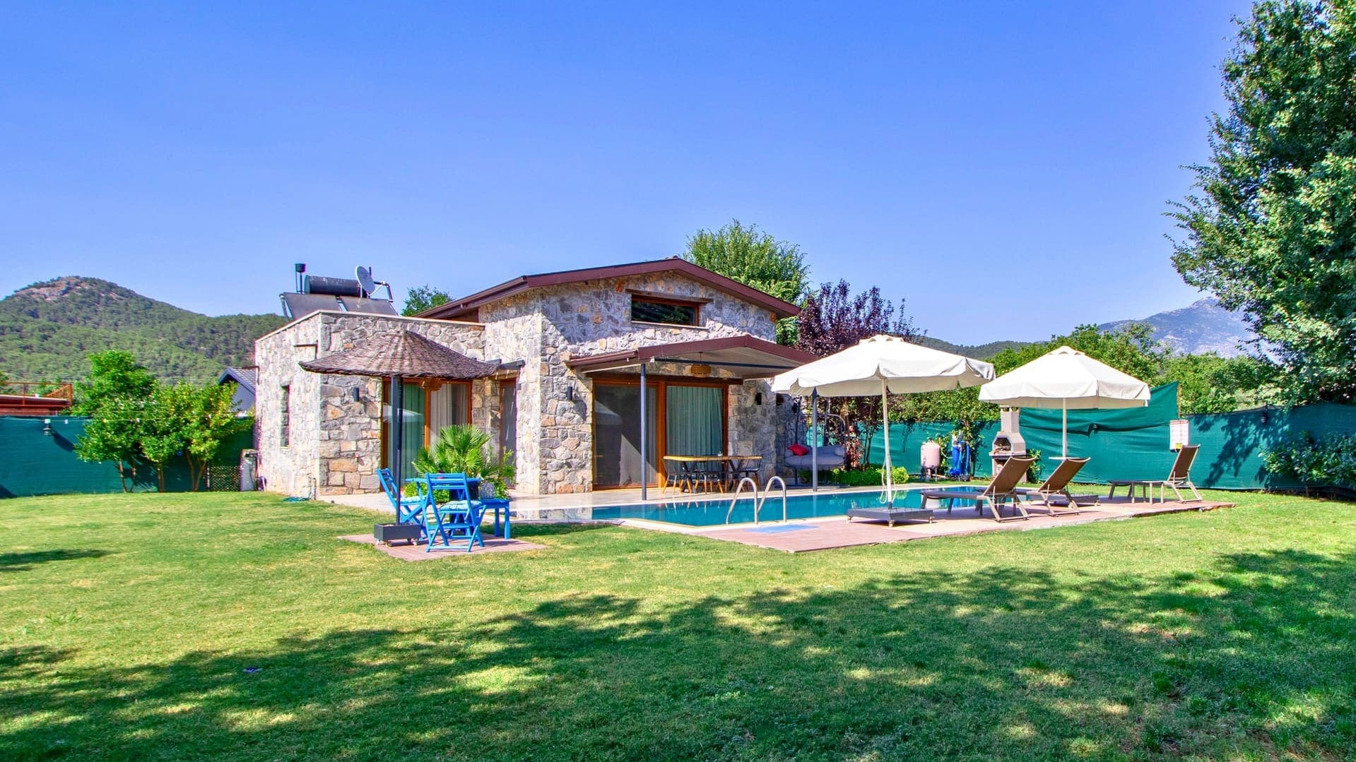 Geniş çim bahçeli, taş cepheli villa ve önünde havuzu.