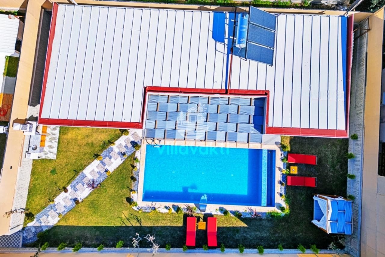 Metal çatılı villanın havuzu, kırmızı şezlongları ve çim alanı.