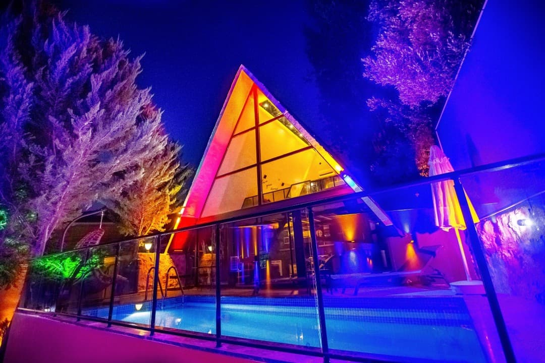 Gece vakti renkli ışıklarla aydınlatılmış üçgen çatılı villa ve önündeki havuz.