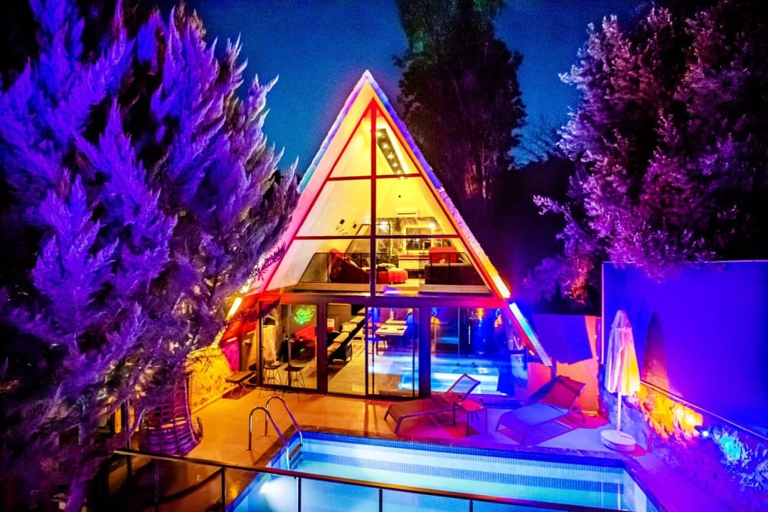 Gece mor ışıklarla aydınlatılmış üçgen çatılı villanın havuzlu bahçesi.