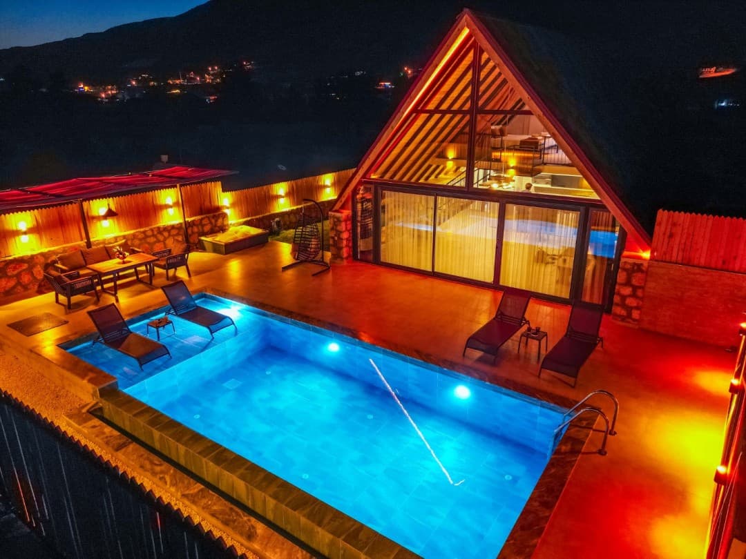 Gece vakti aydınlatmalı havuzu ve şezlongları bulunan üçgen çatılı villa.