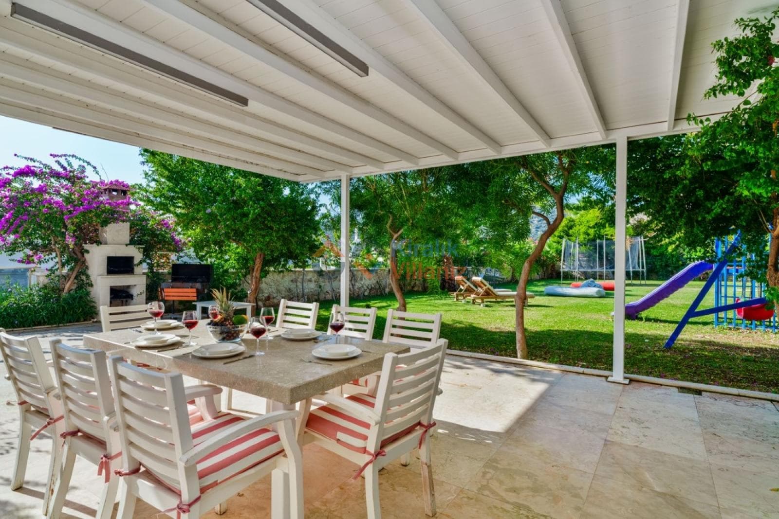 Verandada yemek masası ve sandalyeler, arka planda çimenlik bahçe ve oyun parkı var.