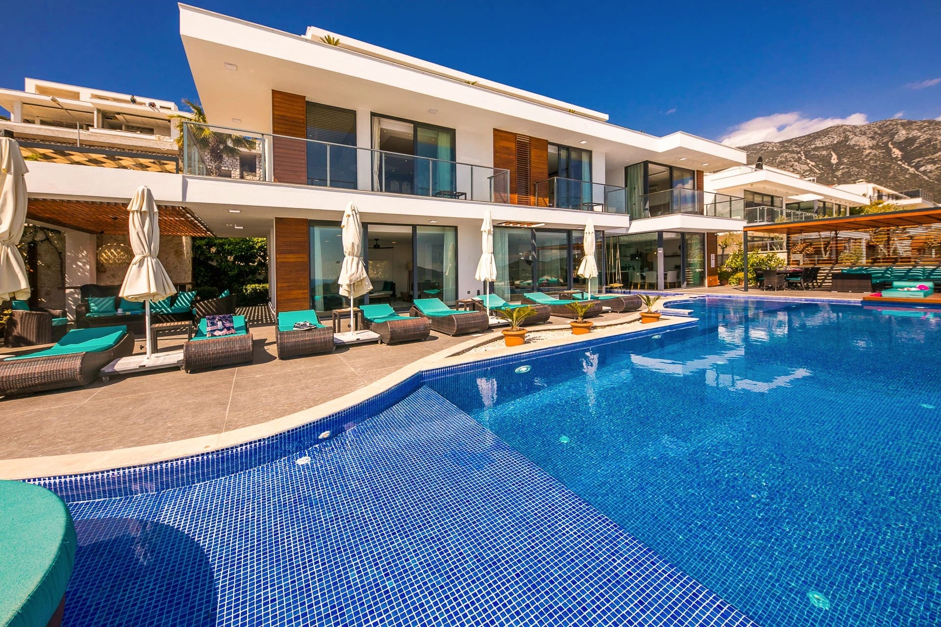 Güneşli günde önünde büyük havuz ve şezlonglar bulunan modern villa.