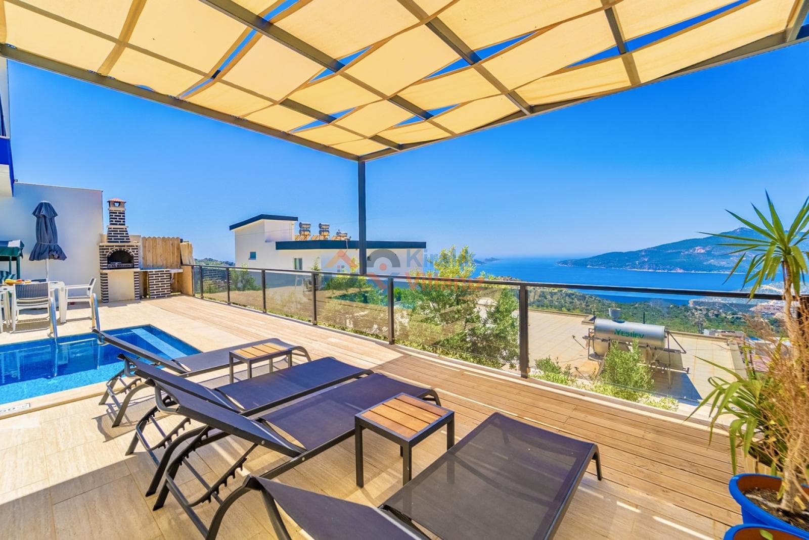 Güneşlikli terasta şezlonglar, barbekü ve deniz manzarası.