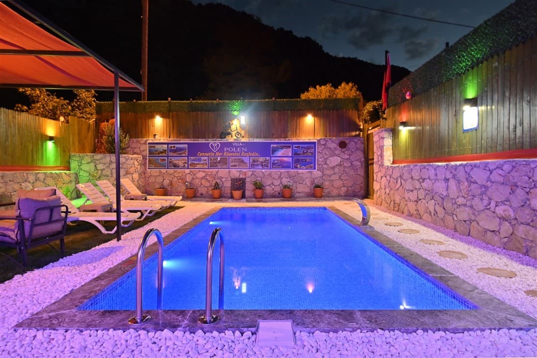 Gece aydınlatmalı havuz, şezlonglar, taş duvar ve üzerinde Villa Polen yazılı tabela bulunan bahçe.