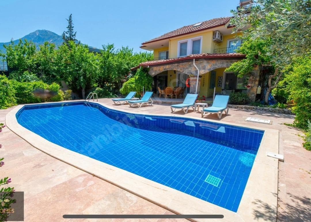 Önünde mavi yüzme havuzu ve şezlonglar bulunan, taş detaylı sarı dış cepheli iki katlı müstakil villa.