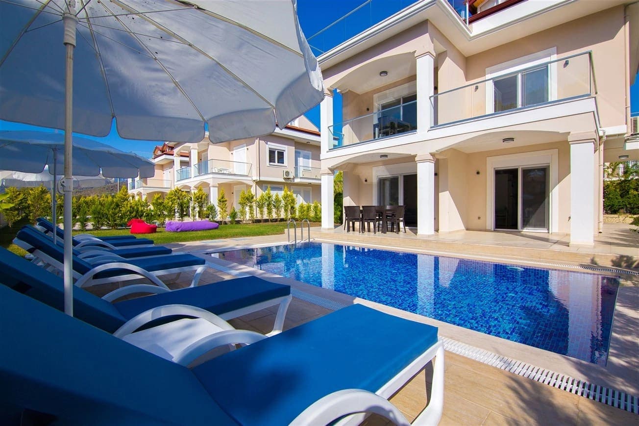 Mavi şezlonglu havuz ve şemsiyeler önünde iki katlı, balkonlu villa.