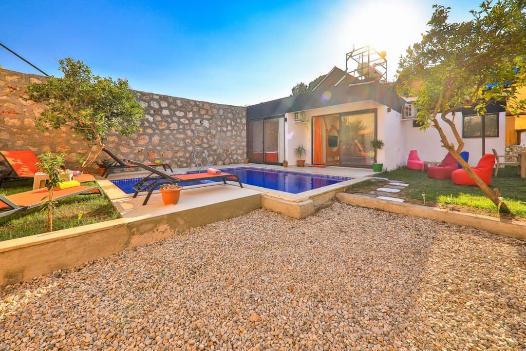Taş duvarlı bahçede havuz, şezlonglar ve arka planda tek katlı beyaz villa.