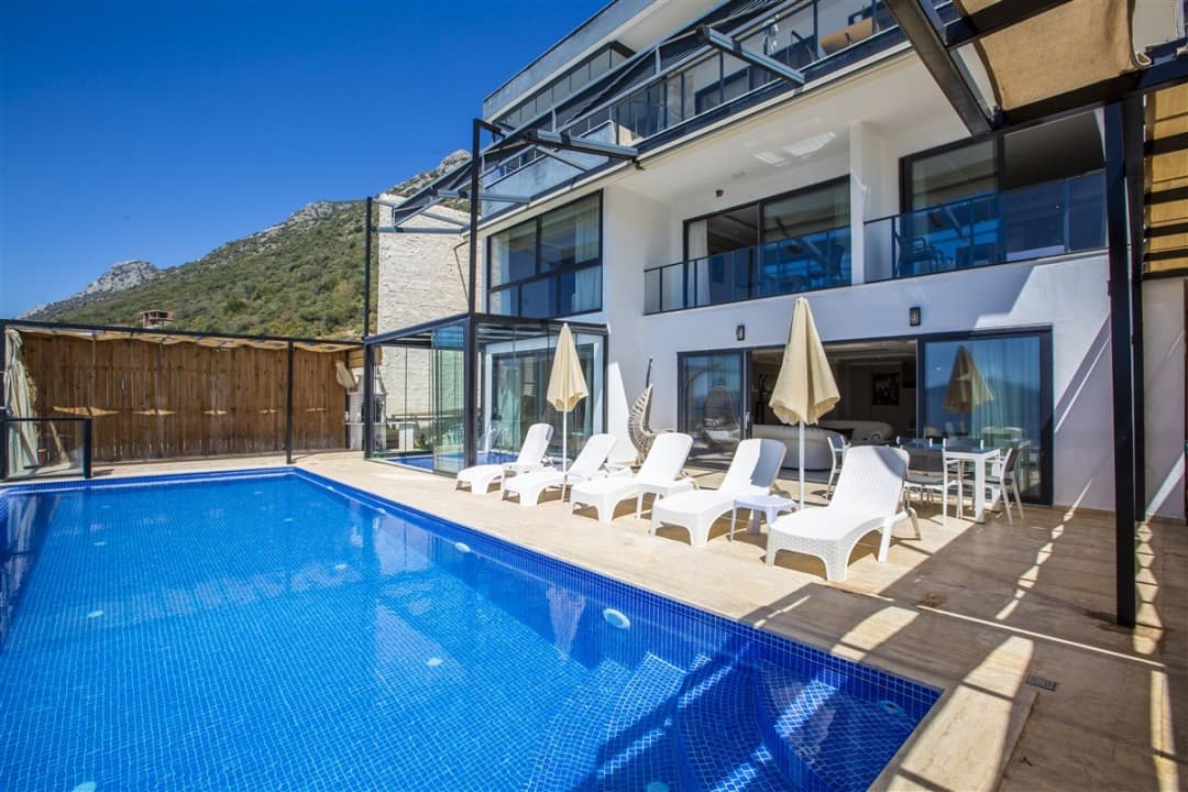 Önünde güneş şezlongları ve şemsiyeler bulunan özel yüzme havuzlu, balkonlu modern beyaz villa.