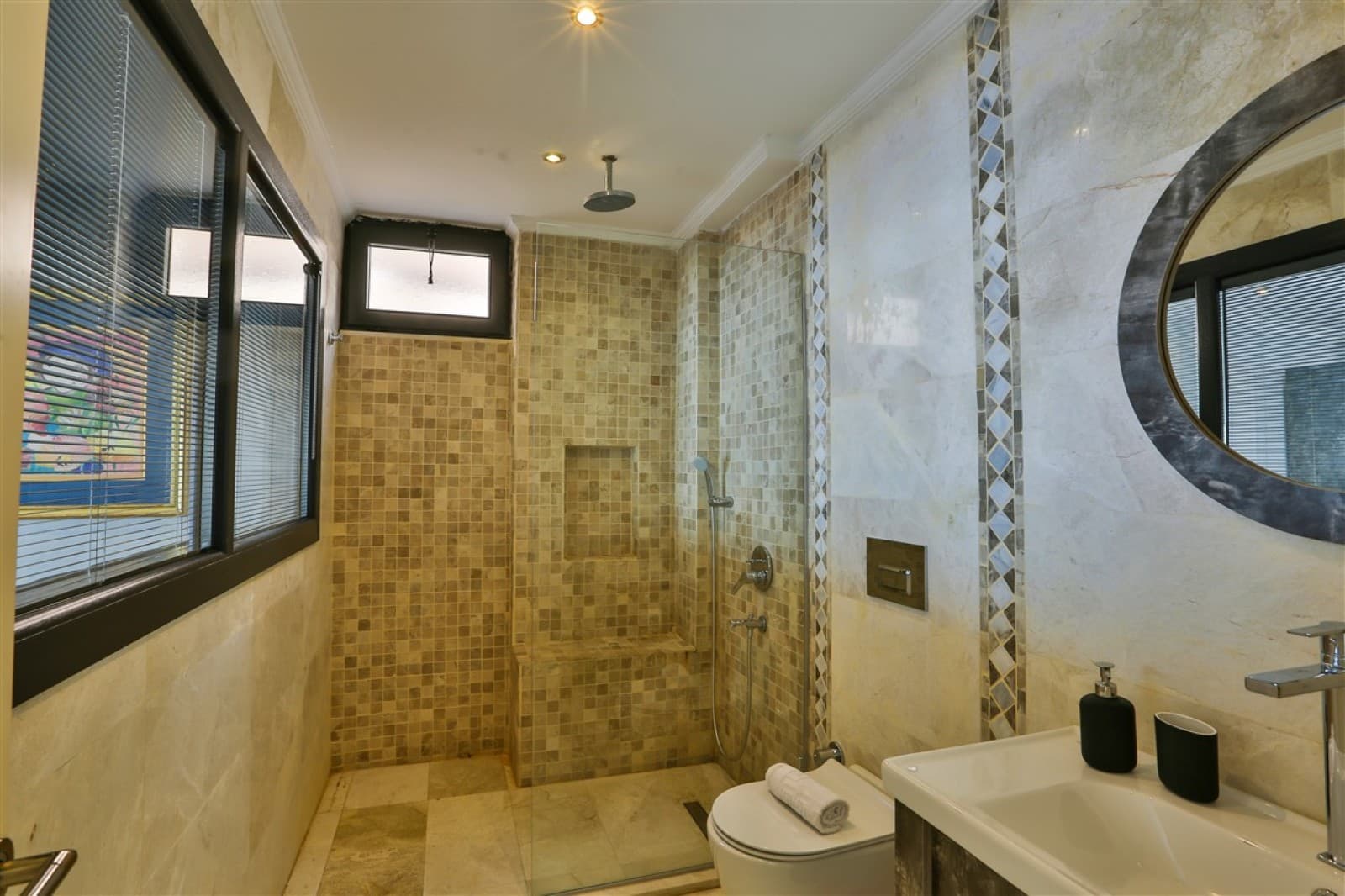 Bej mozaik fayanslı duş alanına sahip modern banyoda lavabo ve tuvalet bulunuyor.
