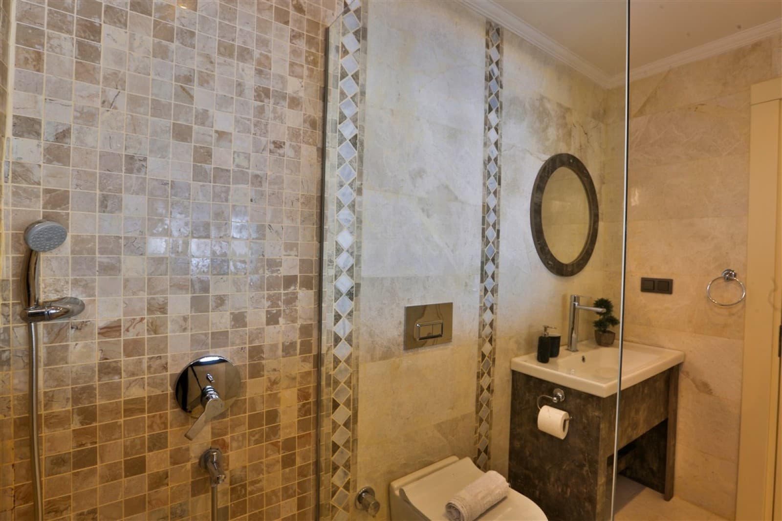 Bej ve kahverengi tonlarında seramik kaplı, cam duşakabinli ve yuvarlak aynalı modern banyo.