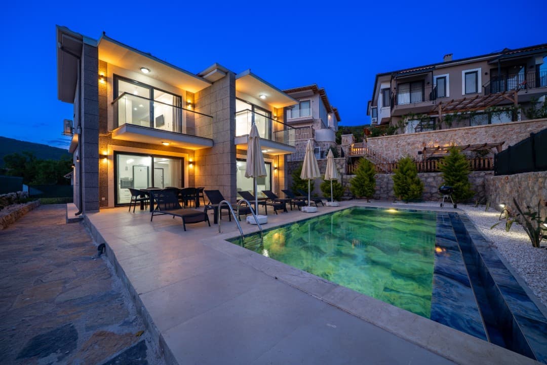 Akşam vakti aydınlatılmış, havuz başında şezlonglar ve yemek masası bulunan iki katlı modern villa.