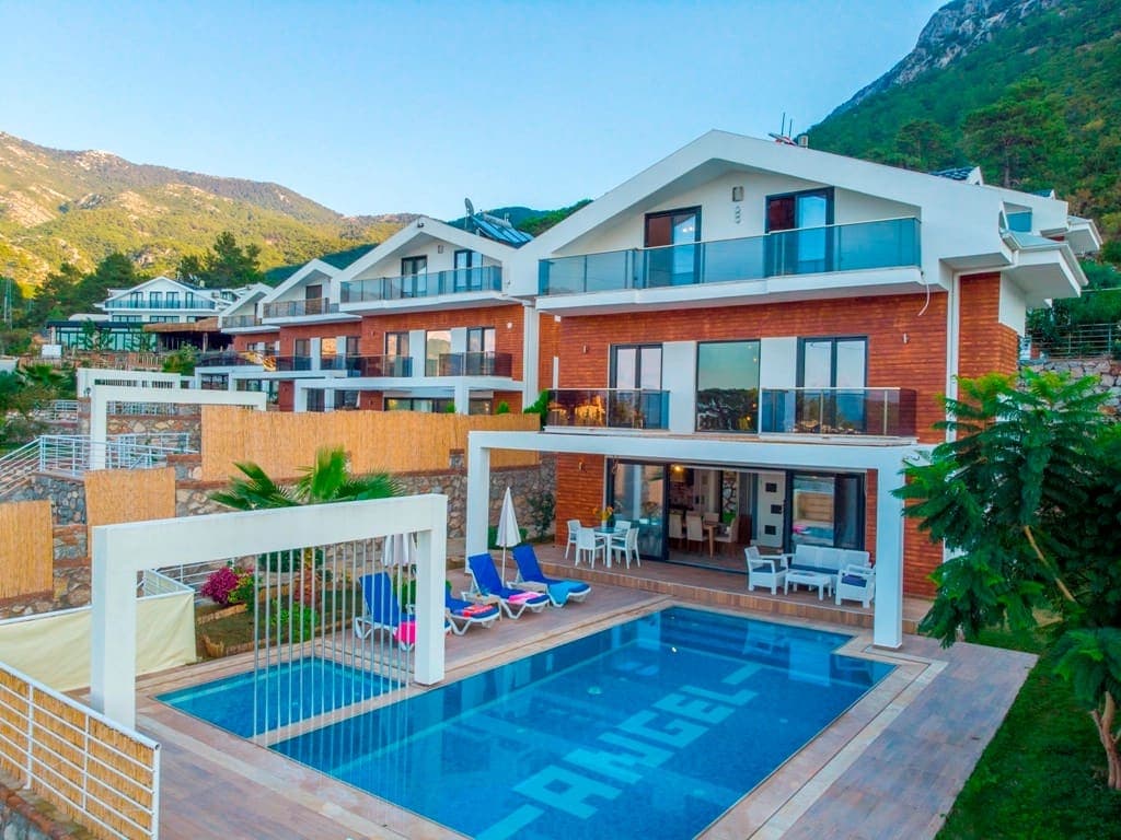 Dağ manzaralı, önünde havuzu ve şezlongları bulunan çok katlı villanın dış cephesi.
